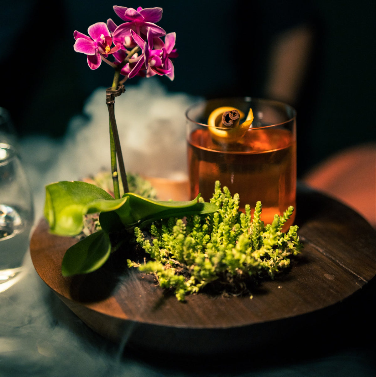 round walnut tray with plant arrangement and drink on it on dark backround surrounded with smoke